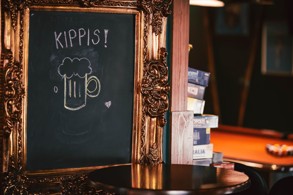 Koristeellisessa kehystetyssä taulussa on liitukirjoitus KIPPIS! ja tuoppipiirros pubin pöydän vieressä.